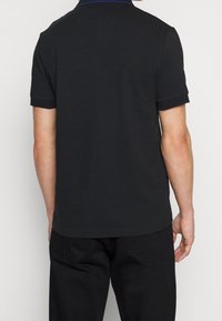 Černé polo tričko s krátkým rukávem, s decentním límečkem a modrými akcenty. Hladká textura látky; střih zobrazen ze zadní strany s viditelnými kalhotami.