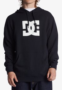Sudadera negra con bolsillo frontal, capucha con cordón y un gran logotipo en blanco de DC con estrella en el pecho. Tejido de mezcla de algodón.
