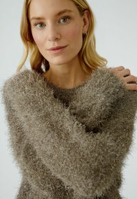 Pull en maille oversized confortable en brun clair, en matière pelucheuse, avec un col large et des manches longues, présentant une surface douce et texturée.