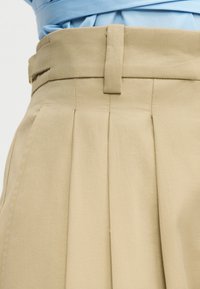 Κοντινή λήψη μπεζ πλεκτών παντελονιών ψηλής μέσης με θηλιές για ζώνη, φορεμένα με γαλάζιο πουκάμισο που έχει μπει μέσα.