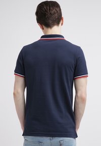 Granatowa koszulka polo z żebrowanym kołnierzykiem, zdobionym czerwonymi i białymi akcentami. Krótkie rękawy z pasującymi prążkowanymi mankietami, wykonana z gładkiego materiału.
