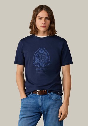 Jeune homme aux cheveux mi-longs portant un t-shirt bleu marine avec le logo Hackett London, un jean bleu et une ceinture marron, debout avec une main dans la poche.