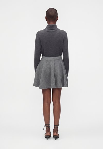 Personne debout de dos portant un pull à col roulé gris foncé, une jupe évasée gris clair et des chaussures noires à talons hauts à brides.