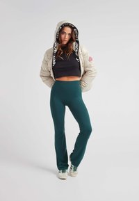 Cappotto imbottito crema con rifiniture in logo nero, crop top nero e leggings verdi scuri a vita alta. Le sneakers verdi e bianche completano il look.