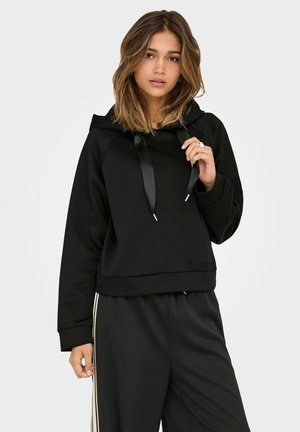 Sudadera negra corta con capucha, con cordones de satén. Hecha de un tejido suave, combinada con pantalones negros que tienen franjas laterales blancas.