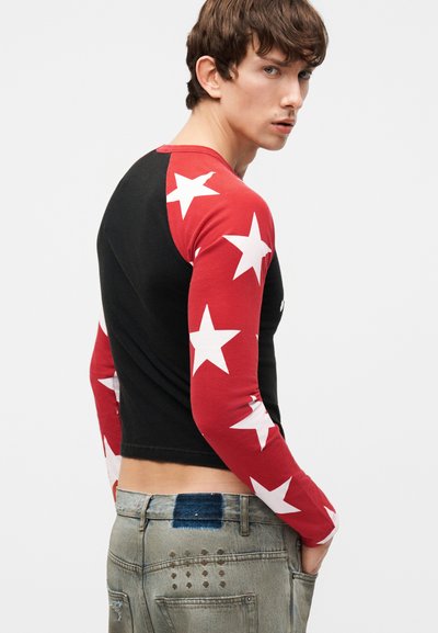 Mladý muž v čiernej košeli s červenými rukávmi zdobenými veľkými bielymi hviezdami, v vybledlých džínsoch, mierne otočený do strany.