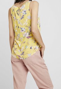 Kukkasonkku ilman hihoja, kirkkaankeltaista kangasta, jossa on violetteja ja valkoisia kuvioita; takana on avainaukko ja kaareva helma.