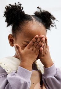 Bambino con capelli ricci e scuri raccolti in due puff, che si copre gli occhi con le mani. Indossa un maglione lavanda con una camicia bianca dal colletto di pizzo.