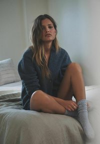 Sininen neulecardigan, jossa on ribbettikaulus ja yksi nappi, käytetty tumman mekon päällä, yhdistetty harmaisiin ribbettisukkiin vaalean värisen vuodevaatteen päällä.