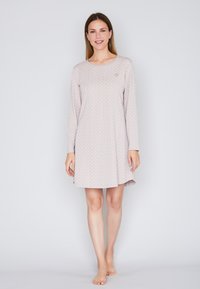 Una donna sta in piedi a piedi nudi indossando un vestito da notte a maniche lunghe di un rosa chiaro con un piccolo motivo geometrico bianco, su uno sfondo semplice e chiaro.
