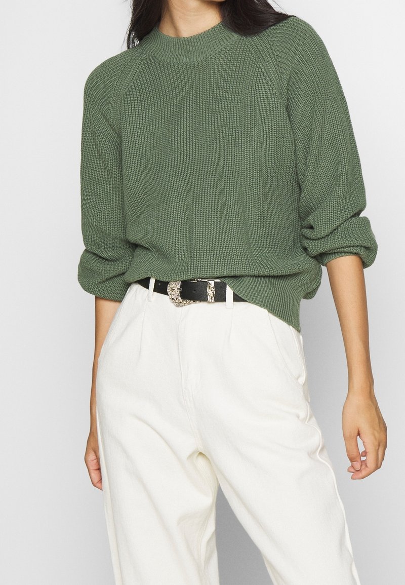 Femme portant un pull vert texturé aux manches retroussées, rentré dans un pantalon blanc taille haute avec ceinture noire et boucle décorative.