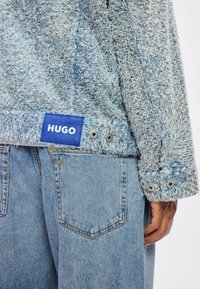Light blue textured sweater with a blue "HUGO" label worn with faded blue denim jeans, showing back pocket and wrist details.