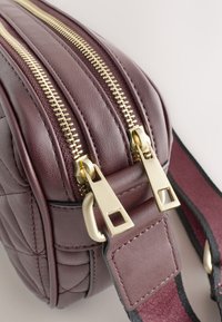 Bordeauxrød lædertaske med quiltet design, udstyret med to guldlynlåse, rektangulære beslag og en tekstureret rem med røde accenter.