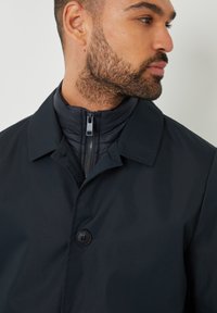 Marineblå frakke med en glat tekstur, udstyret med en høj krave, skjult lynlås og en sort knap til lukning.