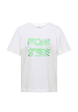 Femme portant un t-shirt blanc avec un motif abstrait vert clair de jardin côtier et une casquette beige, sur fond uni.