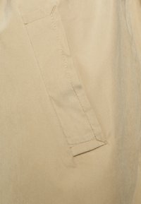 Be�żowa tkanina z boczną kieszenią, wykończoną prostym szwem, o gładkiej fakturze. Kieszeń ma prostokątny kształt i stonowany design.