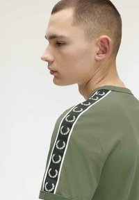 Jeune homme aux cheveux courts portant une chemise vert olive ornée de bandes à motifs de couronne de laurier noire et blanche sur l'épaule.