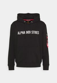 Sudadera negra con la impresión "ALPHA INDUSTRIES" en blanco desgastado, que cuenta con un bolsillo frontal y un bolsillo con cremallera en la manga con un detalle de etiqueta roja.