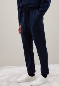 Marineblaue Jogginghose mit einem Kordelzugbund, Seitentaschen und schmal zulaufenden Bündchen, kombiniert mit weißen Sneakers auf einer strukturierten Oberfläche.