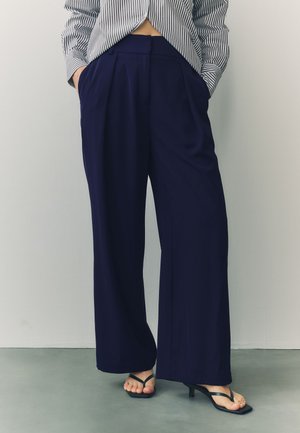 Person trägt hoch taillierte, weit geschnittene marineblaue Hose und schwarze Sandalen mit Absatz, Hände in den Taschen, kombiniert mit einem kurz geschnittenen gestreiften Hemd.