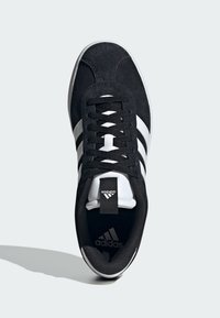 Černé semišové tenisky s bílými třemi pruhy, plochými tkaničkami, polstrovaným límcem a černým vnitřkem. Logo Adidas na jazyku a na boku.