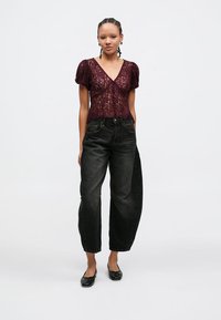Top de renda borgonha com mangas curtas bufantes, combinado com jeans pretos de cintura alta. Modelo usa sapatilhas pretas, em pé sobre um fundo simples.