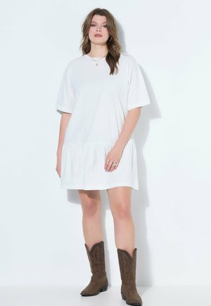 Giovane donna in piedi che indossa un vestito bianco largo a maniche corte e stivali da cowboy marroni ricamati fino al polpaccio su sfondo bianco.