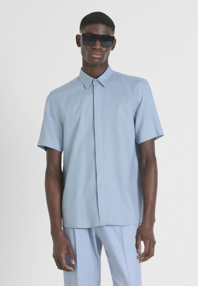 Camicia azzurra chiara a maniche corte con bottoni, realizzata in tessuto testurizzato, abbinata a pantaloni coordinati; modello che indossa occhiali da sole scuri.