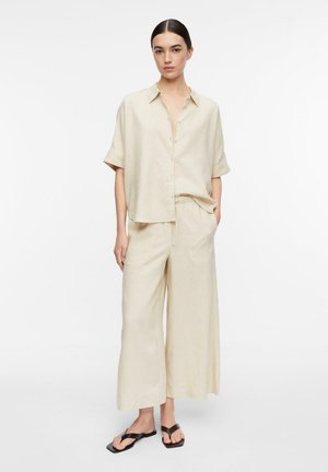 Kobieta stojąca w luźnej beżowej koszuli na krótki rękaw zapinanej na guziki i szerokich spodniach, połączona z czarnymi sandałami typu thong na białym tle.