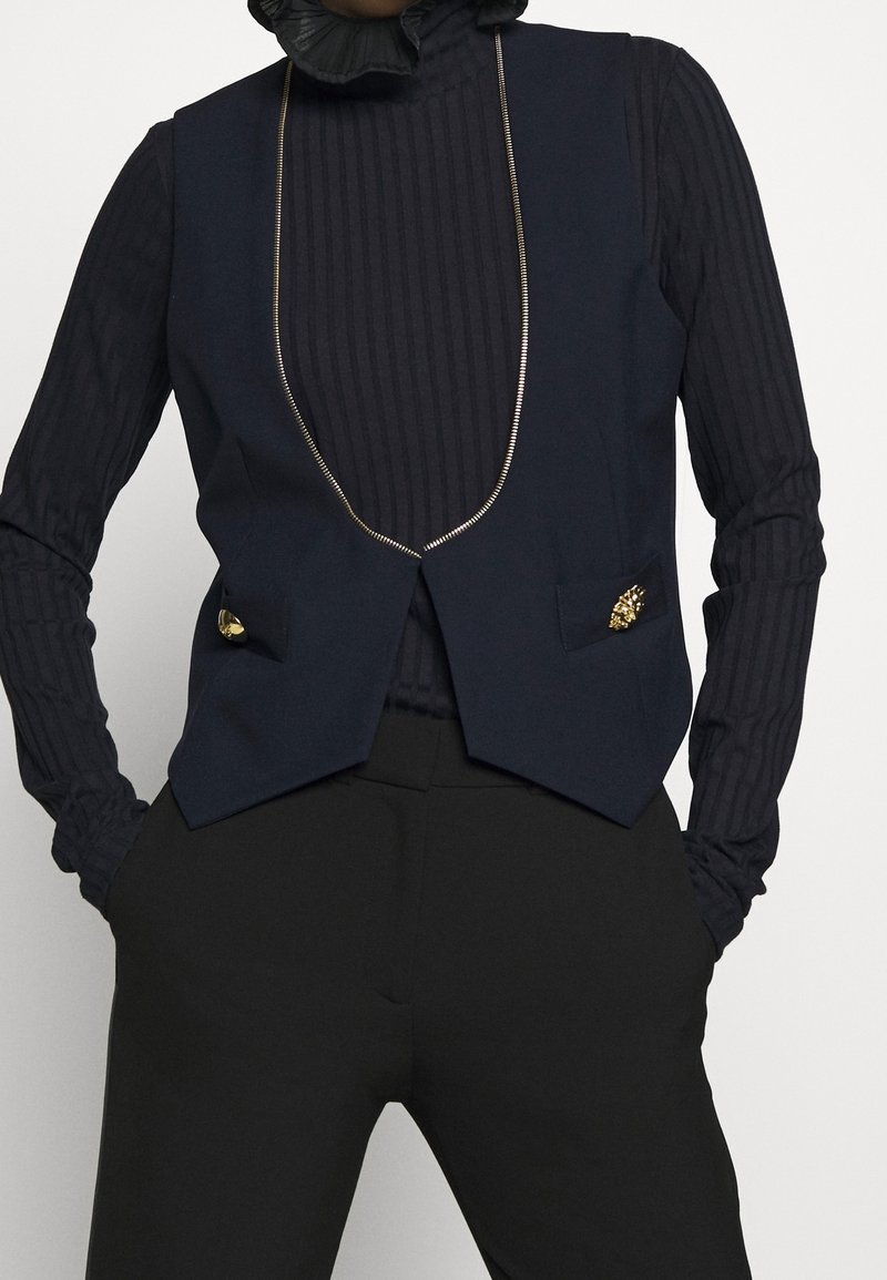 Personne portant un gilet bleu marine à boutons dorés sur un col roulé noir côtelé à manches longues et un pantalon noir, les mains dans les poches.