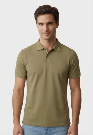 Man die een kort olijfgroen poloshirt en lichtblauwe spijkerbroek draagt, staande tegen een effen witte achtergrond.