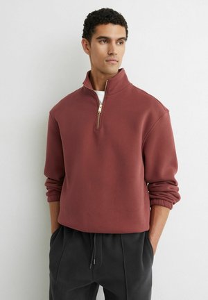 Mees, kes kannab roostekarva kvartstõmblukuga pusa, käised üleskääritud, mustad paeltaskutega püksid, seisab valge seina taustal, käed taskutes.