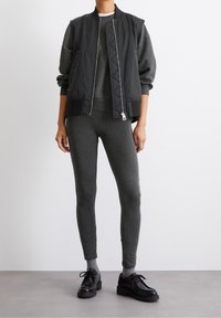 Giacca bomber nera con zip frontale, felpa grigia sotto e leggings grigi aderenti. Scarpe nere con lacci completano l'outfit.