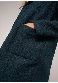 Tummanvihreä teksturoiduista kankaista valmistettu takki, jossa on nappikiinnitys edessä, suuri läppätasku ja pehmeä, pörröinen materiaali.