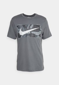 Grå Nike t-shirt med en stor vit swoosh-logotyp över ett rutmönster och en subtil kamouflagebakgrund på framsidan.
