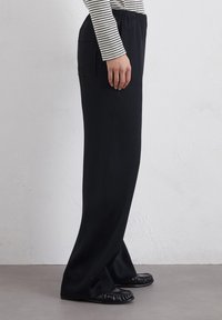 Zwarte lose-fitting broek met een elastische tailleband en zijkleur, gecombineerd met een gestreepte lange mouw top en platte zwarte schoenen.