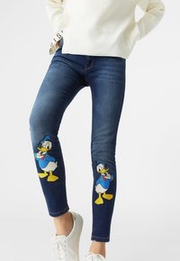Denim jeans in dark blue with two large, colorful Donald Duck appliqués on both knees, paired with a white sweater and white sneakers.