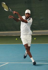 Hvit idretts skjorte med marineblå trim, hvite shorts og en hvit cap. Spilleren svinger en tenniskølle på en blå bane med grønne baller i nærheten.