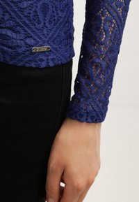 Top lungo a maniche in pizzo blu con motivi intricati e etichetta in metallo del marchio Guess, abbinato a pantaloni neri, mano appoggiata sul fianco.