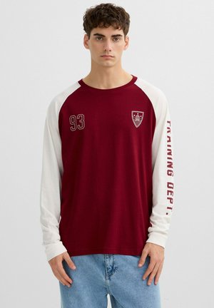 Hombre joven con camiseta de manga larga granate y blanca con el número "93" y un escudo en el frente, texto "TRAINING DEPT." en la manga, y pantalones vaqueros azul claro.