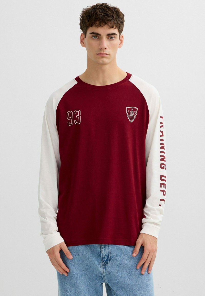 Hombre joven con camiseta de manga larga granate y blanca con el número "93" y un escudo en el frente, texto "TRAINING DEPT." en la manga, y pantalones vaqueros azul claro.