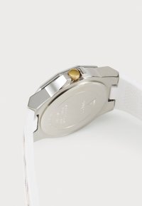 Montre en acier inoxydable argenté avec un boîtier rond, un bracelet en silicone blanc et une couronne en ton or. Le dos présente des détails gravés.