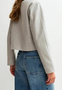 Veste grise courte à manches longues, fabriquée en tissu texturé, associée à un jean bleu taille haute avec des poches arrière.