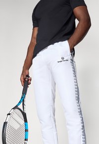 Mand iført hvide Sergio Tacchini træningsbukser med sort logo og holder en sort og blå tennisracket i den ene hånd.