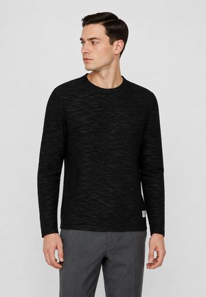Man die een zwarte trui met lange mouwen en textuur draagt en een grijze broek, kijkt naar zijn linkerzijde tegen een effen lichtgrijze achtergrond.