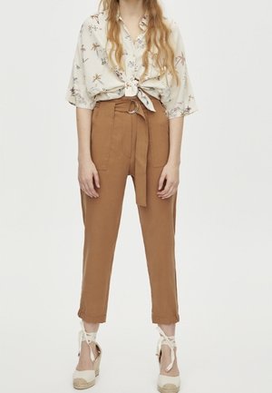 Vrouw draagt hoge taille bruine broek met bindceintuur, bloemenprint korte mouwen blouse geknoopt in de taille en witte espadrille sleehakken met enkelbandjes.