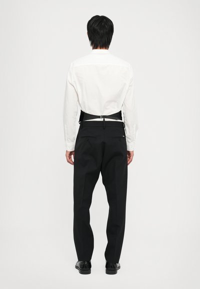 Weißes Langarmhemd mit strukturiertem Rücken und schwarzem Hosenträger. Ergänzt durch schwarze Hose und schwarze Schuhe, was einen formellen Look vermittelt.