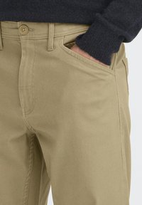 Pantaloni bej din bumbac cu finisaj texturat, prevăzuți cu buzunare frontale și închidere cu un singur nasture în talie. Cusătură laterală vizibilă.