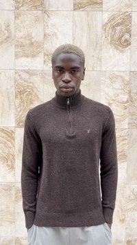 Mladý muž s krátkými blond vlasy, oblečený v tmavě hnědém svršku na zip, stojící proti okrové mramorové obkladačce.