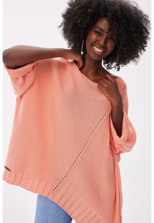 Uśmiechnięta kobieta o kręconych włosach nosi oversize'owy brzoskwiniowy sweter z dzianiny i niebieskie dżinsy, trzymając kołnierz i pozując na białym tle.